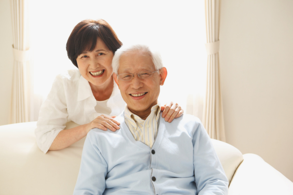 ソファに座る笑顔の高齢男性と、男性の肩に手を添えて笑う高齢女性